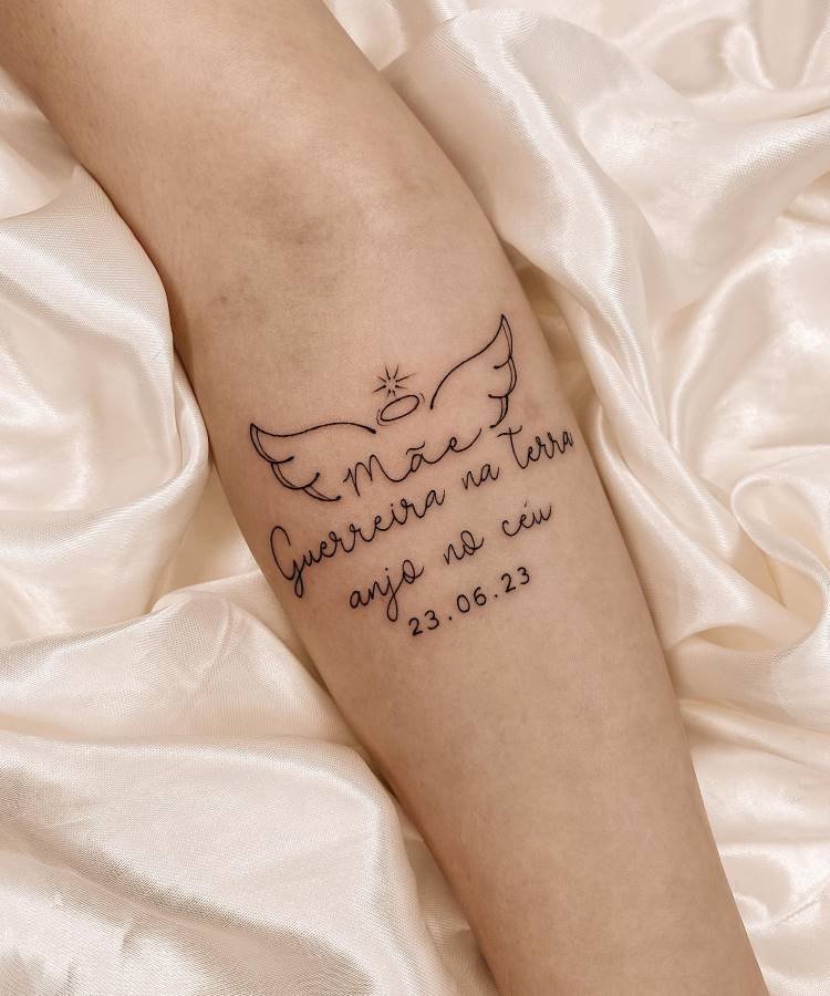 Uma tatuagem delicada no antebraço esquerdo de uma pessoa apresenta a palavra "Mãe" com asas de anjo e uma auréola. Abaixo, as frases "Guerreira na terra" e "anjo no céu" são acompanhadas da data "23.06.23", sugerindo uma homenagem.