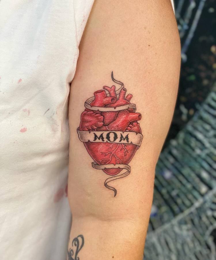 Uma tatuagem vibrante de um coração anatômico em tons de vermelho e rosa está visível no braço. Uma faixa envolve o coração com a palavra "MOM" em letras pretas, demonstrando uma homenagem.