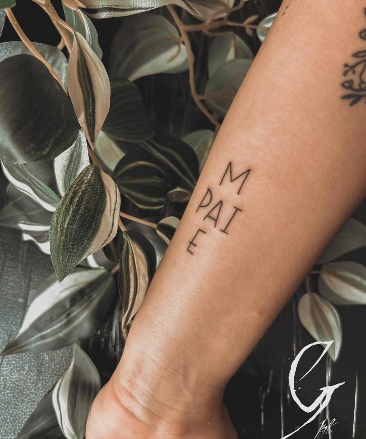 Uma tatuagem minimalista no antebraço, exibindo as palavras "MÃE" e "PAI" verticalmente. Folhas verdes e brancas desfocadas à esquerda e uma tatuagem parcial de uma letra "G" ou "C" na parte inferior complementam a composição.