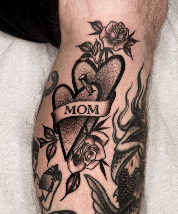 mae-pai-tatuagem-35 A imagem exibe uma tatuagem no estilo "old school" na perna de uma pessoa. Ela apresenta dois corações texturizados, um deles transpassado por um prego, e uma faixa com a palavra "MOM". Rosas com folhas adornam a parte superior e inferior. Há outras tatuagens visíveis na mesma perna.