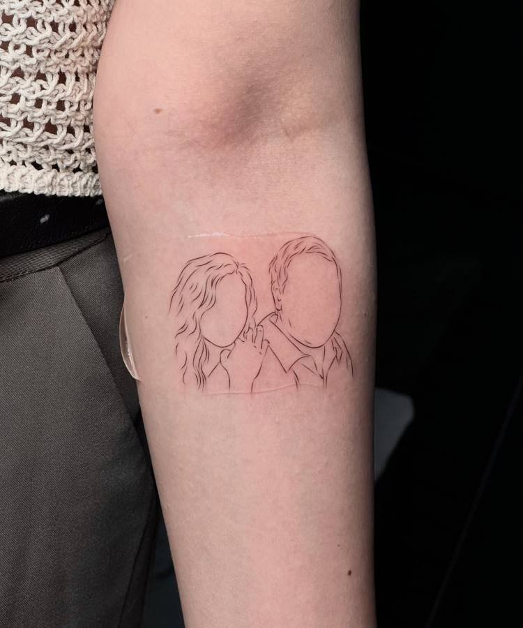 Uma tatuagem de contorno fino no braço exibe as silhuetas de duas pessoas sem traços faciais, uma com cabelo encaracolado e outra com cabelo curto. O desenho minimalista sugere uma conexão pessoal entre os indivíduos.