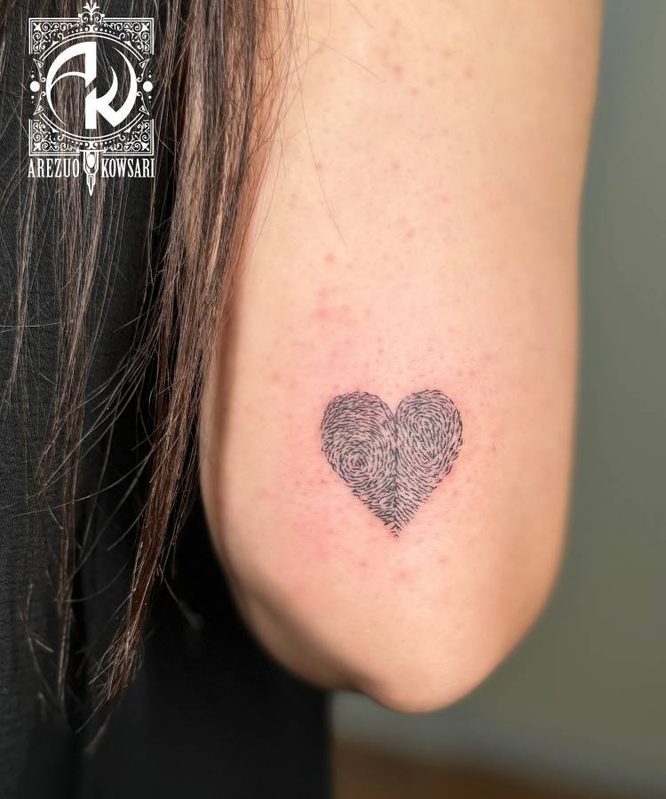 mae-pai-tatuagem-37 A imagem exibe uma pequena tatuagem de coração no braço de uma pessoa. O coração é preenchido com um padrão que se assemelha a uma impressão digital, em tons de cinza escuro. Parte do cabelo escuro da pessoa é visível. No canto superior esquerdo, há um logotipo e o nome "AREZUO KOWSARI".