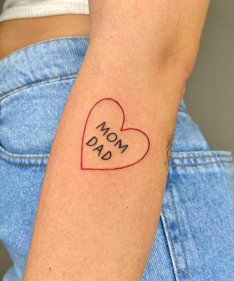 mae-pai-tatuagem-38 A imagem mostra uma tatuagem no braço de uma pessoa. É um contorno de coração em linha fina vermelha, com as palavras "MOM" e "DAD" escritas em preto dentro dele. A pessoa veste calça jeans azul, e a pele é clara.