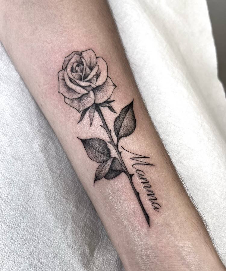 mae-pai-tatuagem-39 A imagem apresenta uma tatuagem em preto e branco de uma rosa com folhas no antebraço de uma pessoa. O caule da rosa se estende para baixo e tem a palavra "Mamma" escrita em itálico ao longo dele. A tatuagem é detalhada e a pele é clara.