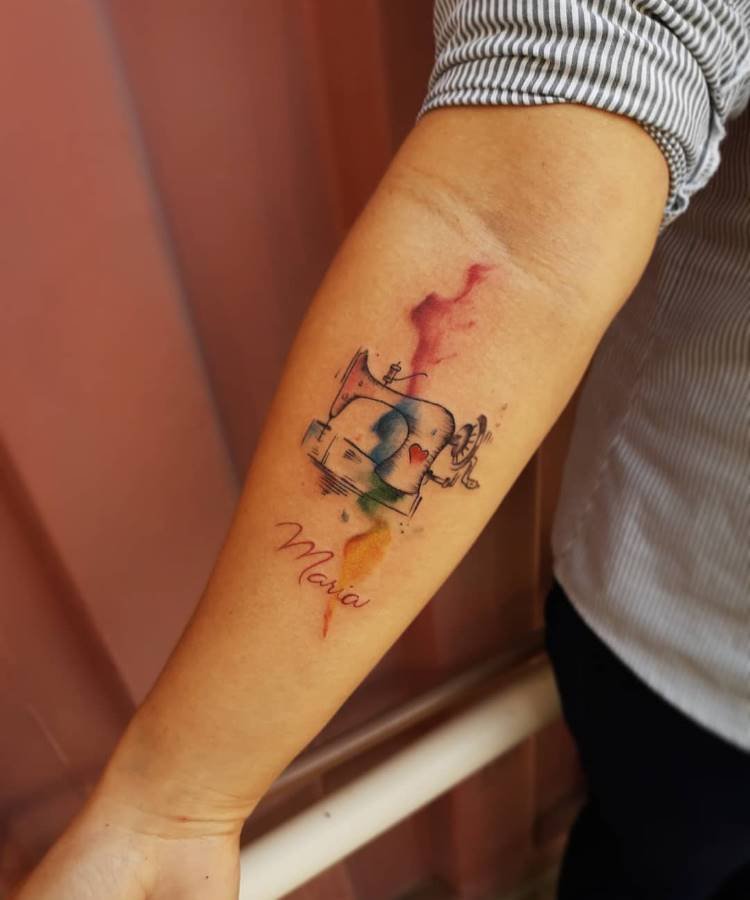 Uma tatuagem vibrante no antebraço de uma pessoa. Apresenta uma máquina de costura em estilo de linha preta sobre manchas de aquarela em tons de vermelho, azul e amarelo. O nome "Maria" está tatuado abaixo do desenho principal.