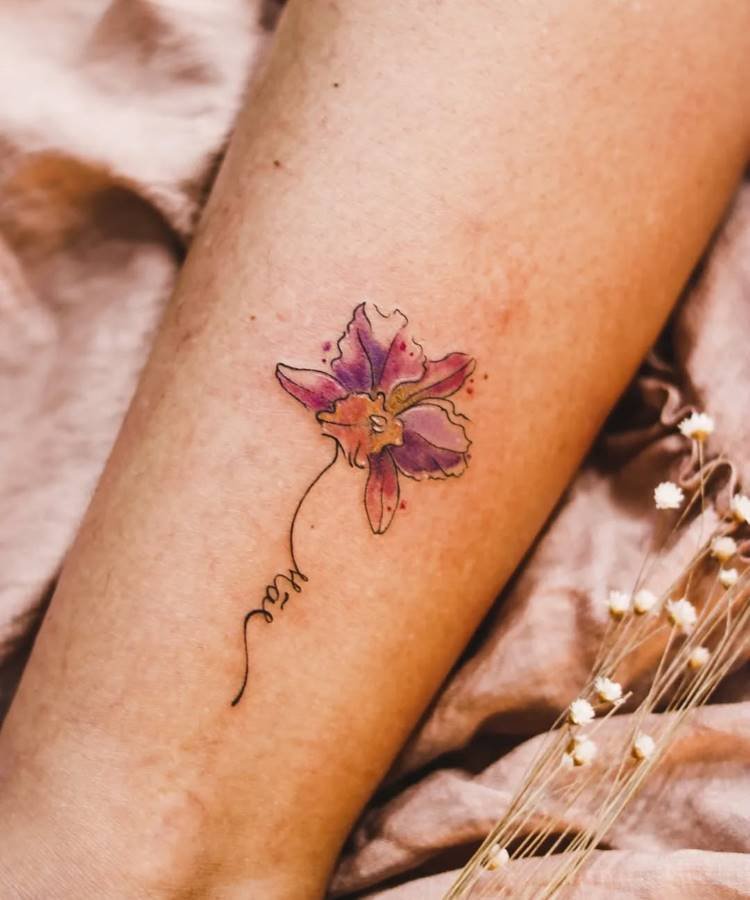 Uma tatuagem delicada de uma orquídea roxa com tons de rosa e detalhes amarelos está no antebraço. O caule da flor se estende e forma a palavra "Mãe" em caligrafia. Pequenos pontos de cor ao redor realçam o design.
