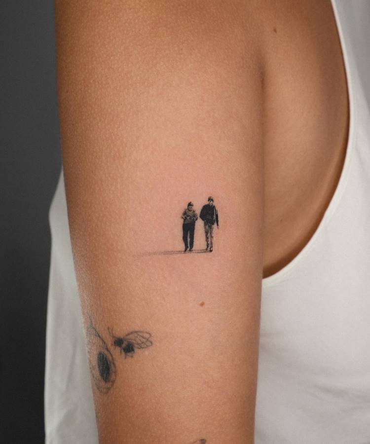 mae-pai-tatuagem-7 A imagem exibe uma pequena tatuagem minimalista no braço de uma pessoa, retratando duas figuras de corpo inteiro, possivelmente masculinas, caminhando lado a lado. A tatuagem é em preto e cinza, com sombras que dão profundidade. Abaixo, há um esboço de outra tatuagem com abelhas e uma flor.