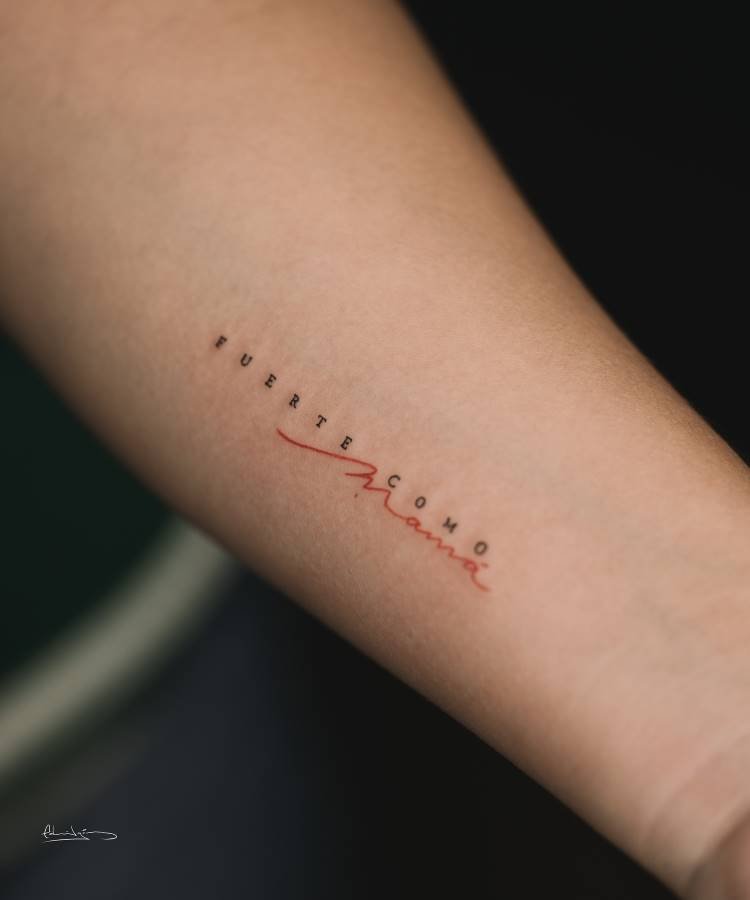 mae-pai-tatuagem-8 A imagem mostra uma tatuagem no antebraço de uma pessoa. A tatuagem diz "FUERTE COMO" em letras minúsculas pretas, e "Mama" em letras cursivas vermelhas, com o "Ma" sublinhado e as letras se estendendo. A pele é clara e o fundo escuro.