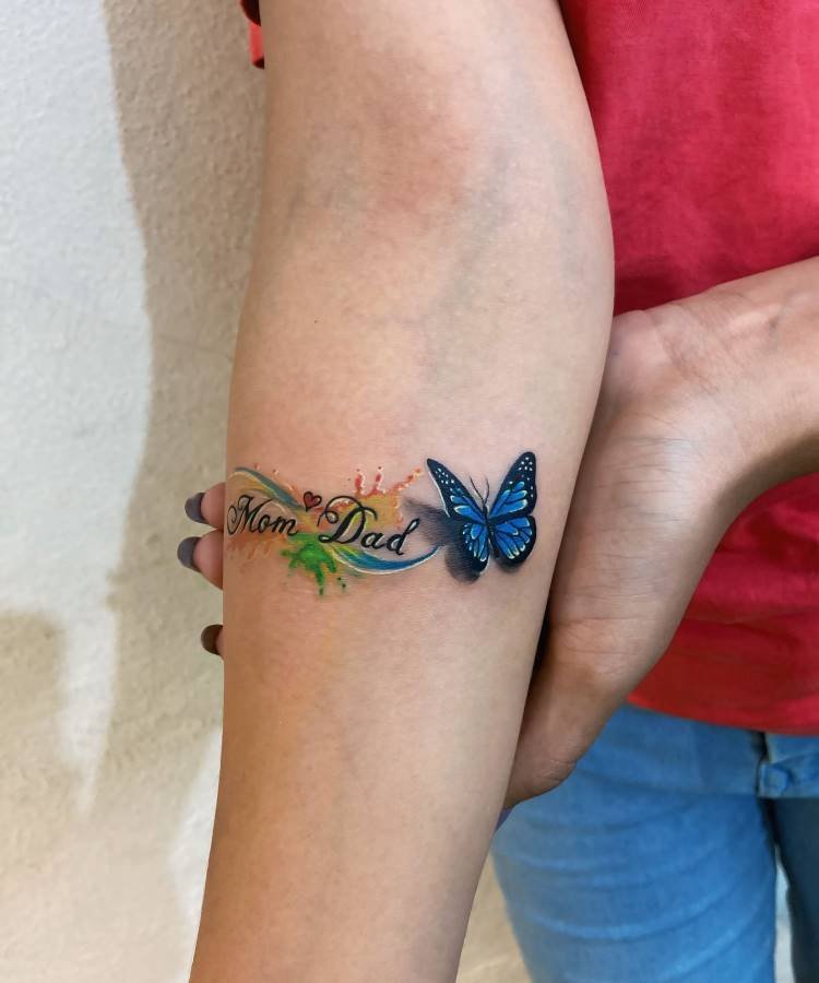 Uma tatuagem vibrante no antebraço esquerdo de uma pessoa. Inclui um fluxo de respingos de tinta verde, amarela e azul, com as palavras "Mom" e "Dad" em destaque. Uma borboleta azul 3D repousa no final da frase.