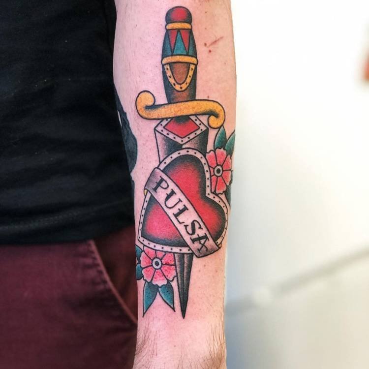 Adaga colorida old school enfiada em coração vermelho com faixa escrito "pulsa" feita no braço de um homem de pele clara usando blusa preta e calça vinho