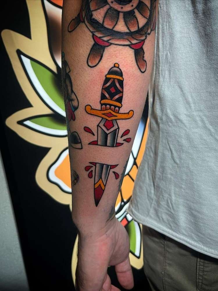 Tatuagem de adaga old school colorida no braço de pessoa de pele clara usando camiseta branca