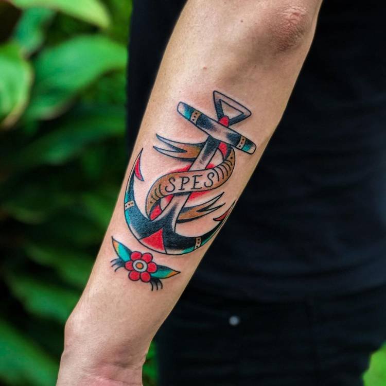 Pessoa de pele clara com âncora azul e vermelha old school tatuada no braço