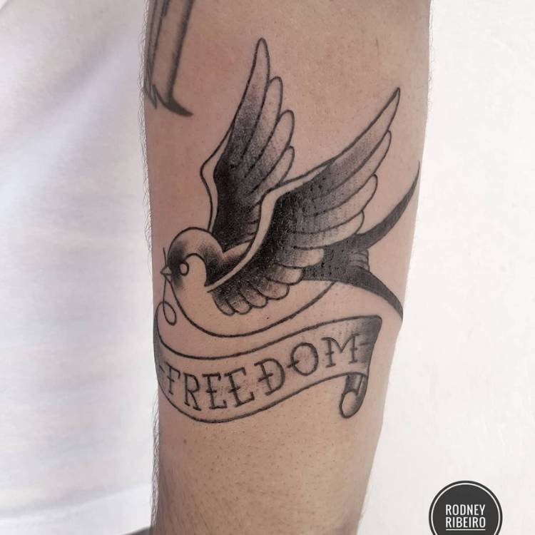 Pessoa de pele clara com tatuagem preta de andorinha old school com faixa escrito "freedom"