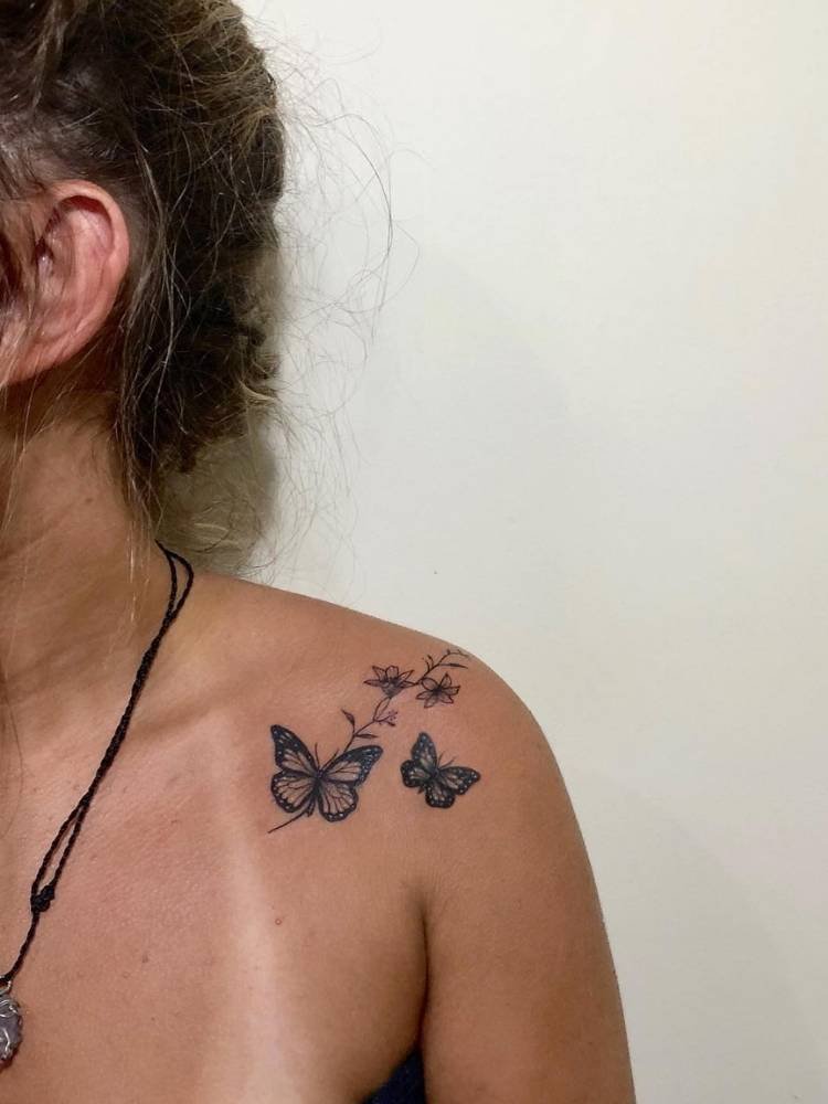 ombro-borboleta-flor Borboletas pequenas em traços finos e um pouco mais grossos com ramos de flores no ombro de mulher de pele bronzeada, com marca da parte superior do biquíni e cabelo marrom preso.