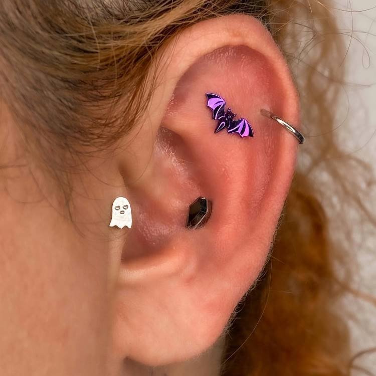 Orelha com piercing anodizado em roxo de morcego na scapha, fantasma anodizado em prata no tragus, argola no hélix e caixão no conch.