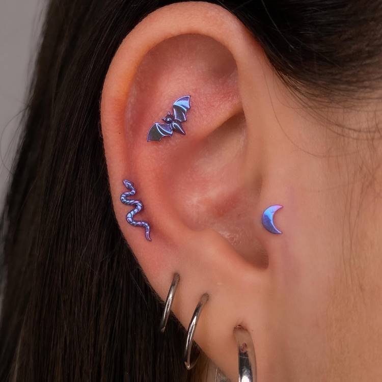 Orelha com morcego em roxo na scapha, cobra em roxo no conch, lua em roxo no tragus e três argolas em prata no lóbulo.