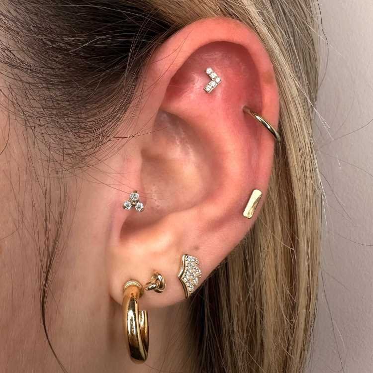 Orelha com piercing de coração na scapha, argola na hélix, piercing no tragus e três brincos no lóbulo.