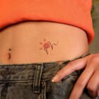 tattoo-cintura-140x140-1-9 A imagem mostra a parte inferior do abdômen de uma pessoa, com um top laranja e jeans. Há um piercing no umbigo e uma tatuagem delicada de flor com traços finos e pétalas vermelhas. A mão está levemente posicionada sobre o cós da calça, destacando o estilo casual e jovem.
