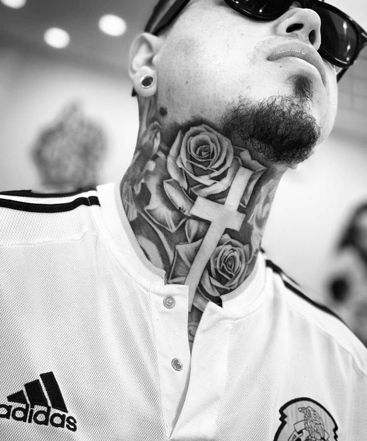 tatuagem-de-cruz-no-pescoco-1-1 A imagem mostra uma pessoa de frente com a cabeça inclinada para trás. Ela tem uma tatuagem no pescoço de grandes rosas e uma cruz central. A pessoa usa óculos escuros, brincos e uma camisa branca da Adidas.
