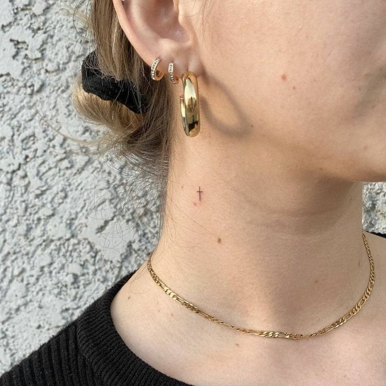 tatuagem-de-cruz-no-pescoco-2-1 A imagem mostra uma pessoa de perfil com cabelo preso e brincos dourados. Ela tem uma tatuagem no pescoço de uma pequena cruz preta, localizada logo abaixo da orelha. A tatuagem é discreta e minimalista. A pessoa também usa uma corrente dourada fina e uma camiseta preta.