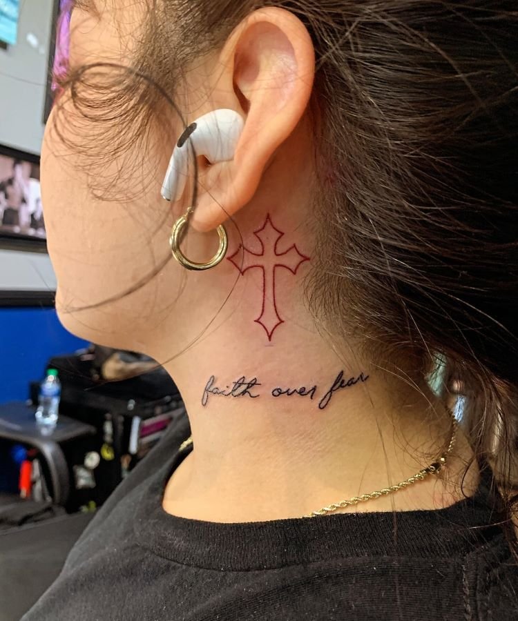 tatuagem-de-cruz-no-pescoco-3-1 A imagem mostra uma pessoa de perfil com cabelo preso. Ela tem uma tatuagem no pescoço de uma cruz vermelha estilizada localizada logo abaixo da orelha. Abaixo da cruz, há uma frase em preto que diz "faith over fear" (fé sobre medo). A pessoa usa um brinco dourado grande e um fone de ouvido.