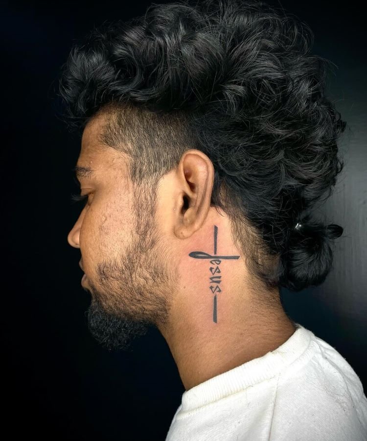 tatuagem-de-cruz-no-pescoco-4-1 A imagem mostra uma pessoa de perfil com cabelo encaracolado preso em um coque. Ela tem uma tatuagem no pescoço que consiste na palavra "Jesus" escrita verticalmente, formando uma cruz. A pessoa usa uma camiseta branca.