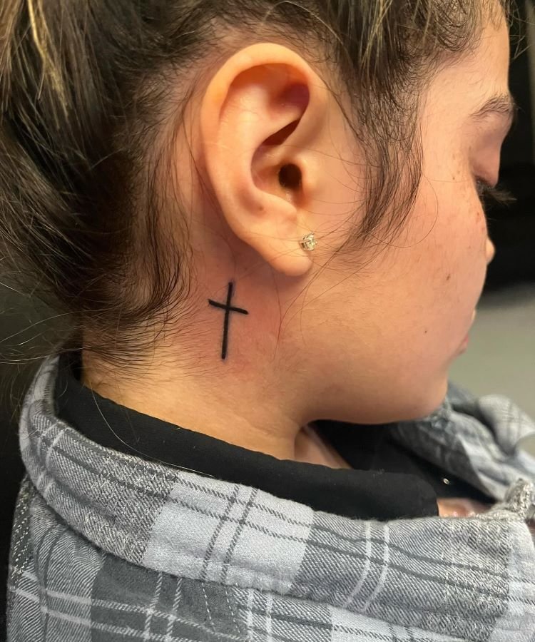 tatuagem-de-cruz-no-pescoco-5-1 A imagem mostra uma pessoa de perfil com cabelo preso. Ela tem uma tatuagem no pescoço de uma pequena cruz preta localizada atrás da orelha. A pessoa usa um brinco pequeno e está vestida com uma camisa xadrez cinza e preta.