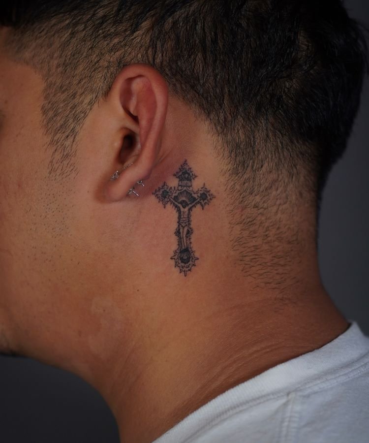 tatuagem-de-cruz-no-pescoco-7-1 A imagem mostra uma pessoa de perfil com cabelo curto e escuro. Ela tem uma tatuagem no pescoço de uma cruz detalhada em preto, localizada logo abaixo da orelha. A pessoa usa uma camiseta branca e pequenos piercings na orelha.