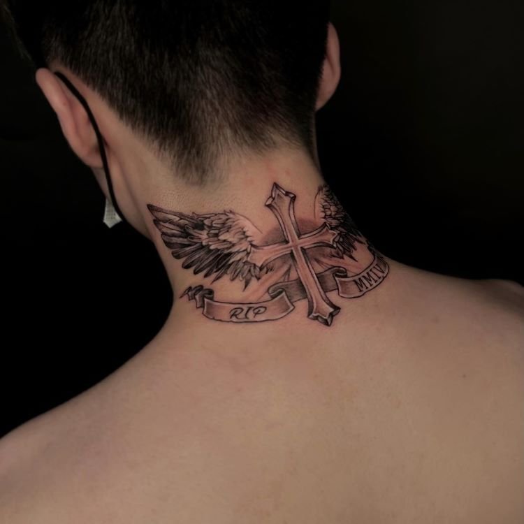 tatuagem-de-cruz-no-pescoco-9-1 A imagem mostra uma pessoa de costas com cabelo curto. Ela tem uma tatuagem no pescoço de uma cruz com asas abertas, detalhada em preto e cinza. Abaixo da cruz, há uma faixa com as inscrições "RIP" e "MMIV". A tatuagem cobre a parte de trás do pescoço até os ombros.