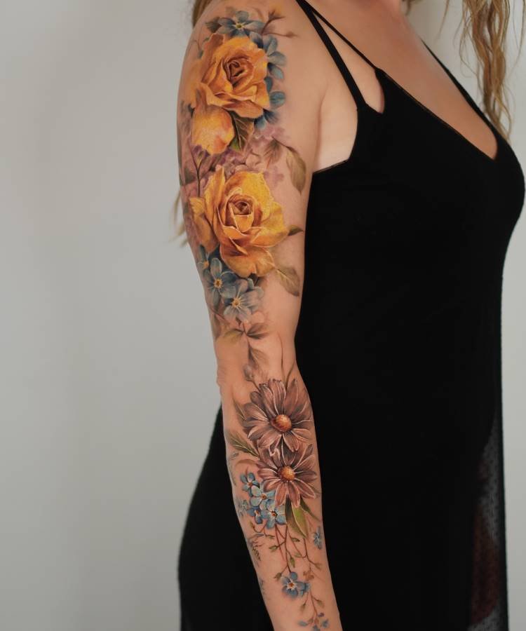 Tatuagem vibrante no braço com rosas amarelas detalhadas, flores marrons e pequenas flores azuis. Os elementos botânicos são realistas, com sombras suaves e folhagens. Ao fundo, uma pessoa com vestido preto e parede de tom claro neutro.