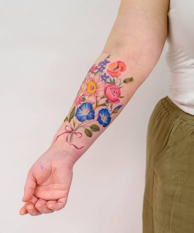 Tatuagem colorida no antebraço com flores detalhadas como rosas, hibiscos, e miosótis em tons vibrantes de azul, rosa e laranja. As flores estão amarradas com uma fita rosa. Fundo neutro com roupa em tons claros e verdes.
