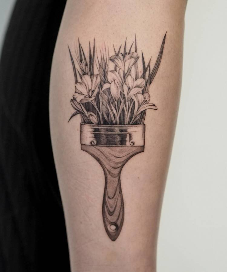 Tatuagem em preto e cinza de um pincel, cuja ponta é composta por flores e folhas delicadas, criando uma ilusão orgânica. Os detalhes realistas destacam a textura do cabo e das flores, que emergem de forma artística.