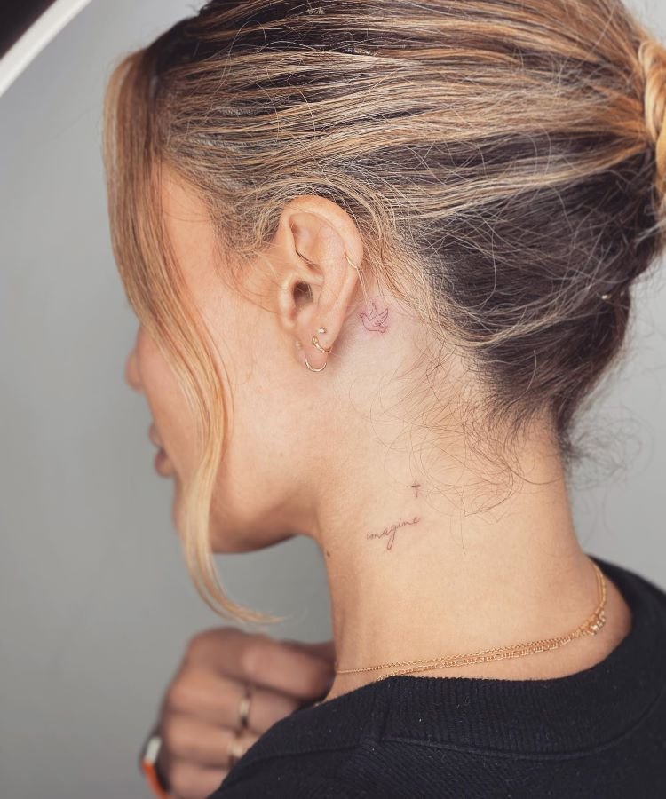 tatuagem-escrita-no-pescoco-1-1 A imagem mostra uma pessoa de perfil com cabelo loiro preso em um coque. Ela tem uma tatuagem no pescoço de uma pequena pombinha atrás da orelha e a palavra "imagine" escrita em vermelho com uma pequena cruz preta. A pessoa usa brincos pequenos e uma corrente dourada.