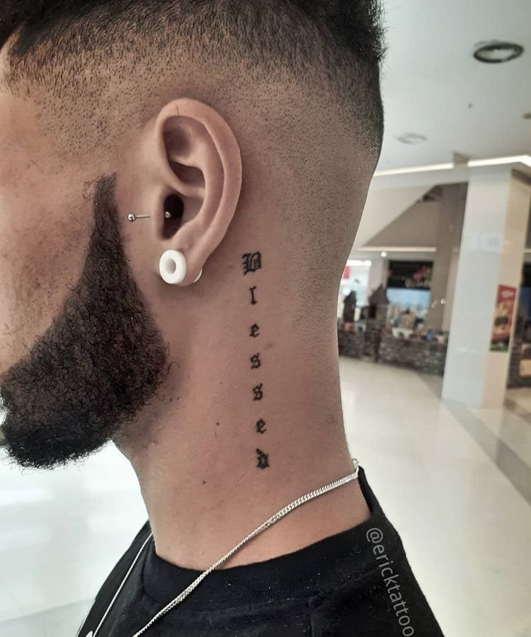 tatuagem-escrita-no-pescoco-4-1 A imagem mostra uma pessoa de perfil com cabelo curto e barba. Ela tem uma tatuagem no pescoço com a palavra "Blessed" escrita verticalmente em letras pretas. A tatuagem está localizada na lateral do pescoço, perto da orelha. A pessoa também usa um alargador branco na orelha e um brinco de argola.
