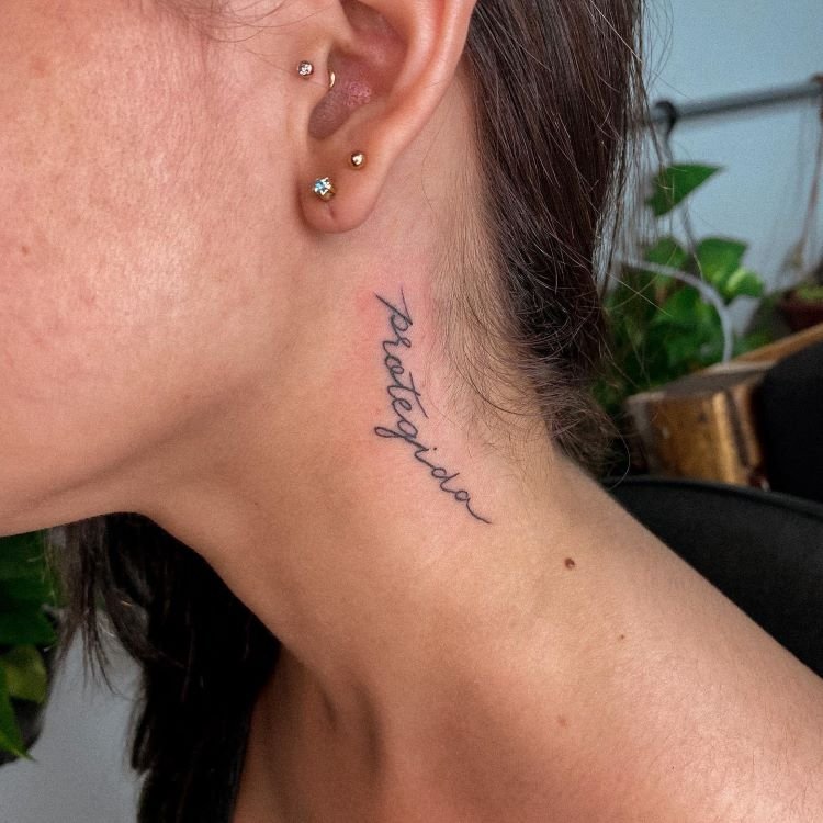 tatuagem-escrita-no-pescoco-7-1 A imagem mostra uma pessoa de perfil com cabelo preso. Ela tem uma tatuagem no pescoço com a palavra "protegida" escrita em letras cursivas pretas. A tatuagem está localizada na lateral do pescoço, perto da orelha, onde a pessoa também usa vários brincos pequenos.