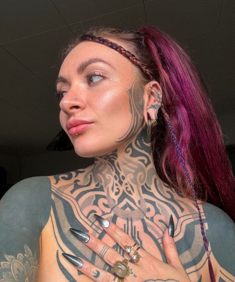 tatuagem-feminina-no-pescoco-11-1 A imagem mostra uma pessoa de perfil com cabelo roxo preso em um rabo de cavalo. Ela tem uma tatuagem no pescoço e no peito de padrões tribais em preto, que se estendem até o rosto. A tatuagem é complexa e cobre grande parte da pele visível.