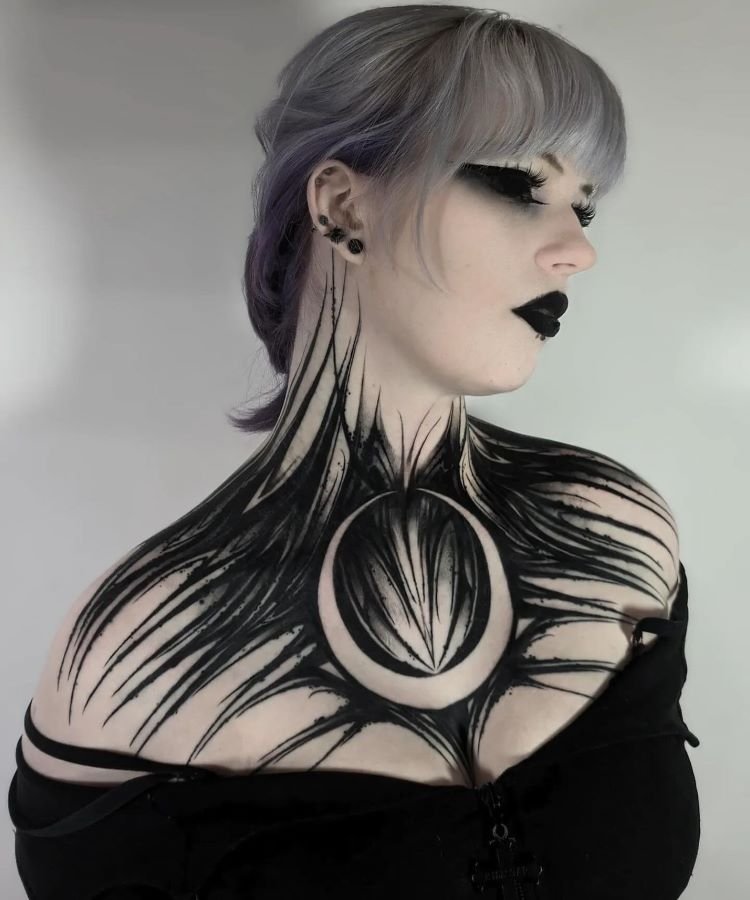 tatuagem-feminina-no-pescoco-13-1 A imagem mostra uma pessoa com cabelo cinza claro e maquiagem escura. Ela tem uma tatuagem no pescoço que se estende até o peito, com um design abstrato em preto, lembrando penas ou garras, e um círculo central formando uma lua. A tatuagem cobre o pescoço e a parte superior do peito.