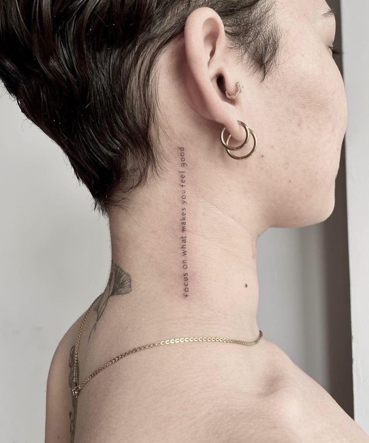 tatuagem-feminina-no-pescoco-17-1 A imagem mostra uma pessoa de perfil com uma tatuagem no pescoço que consiste em uma frase escrita verticalmente. A frase diz "focus on what makes you feel good". A pessoa tem cabelo curto, usa dois brincos de argola dourados e uma corrente dourada no pescoço.