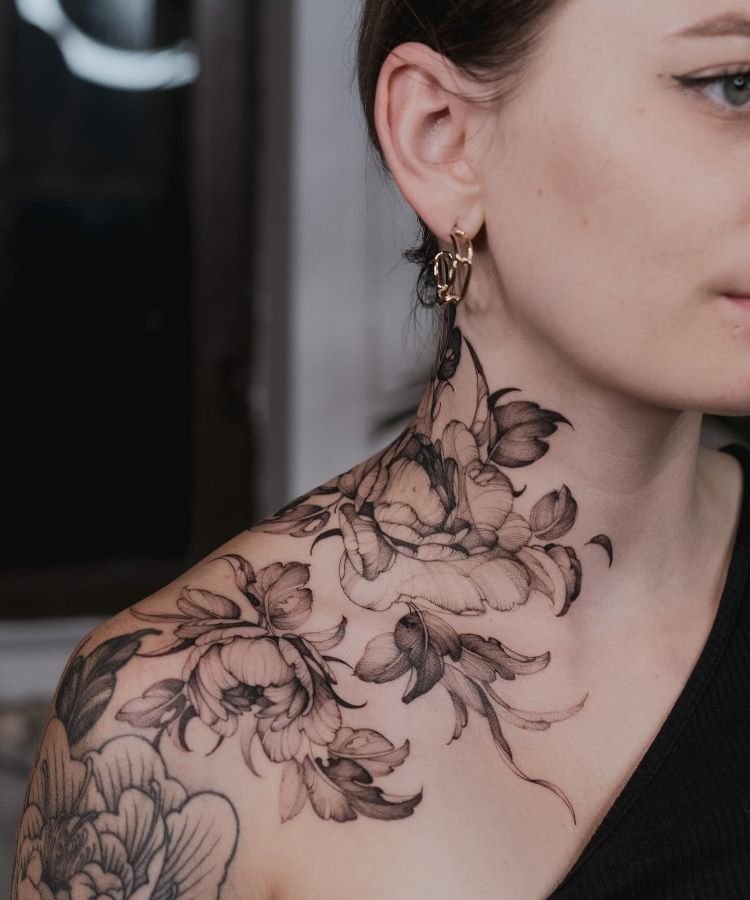 tatuagem-feminina-no-pescoco-18-1 A imagem mostra uma pessoa de perfil com cabelo castanho preso. Ela tem uma tatuagem no pescoço de flores detalhadas em preto e cinza, que se estendem até o ombro. A tatuagem é delicada e realista, cobrindo a lateral do pescoço e parte do ombro.