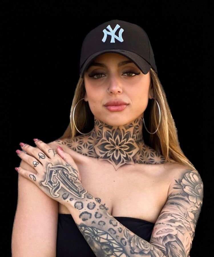 tatuagem-feminina-no-pescoco-19-1 A imagem mostra uma pessoa de frente com cabelo loiro, usando um boné preto e brincos grandes. Ela tem uma tatuagem no pescoço de um grande padrão geométrico em preto, que se estende até o peito. O braço direito também está coberto por tatuagens intricadas.