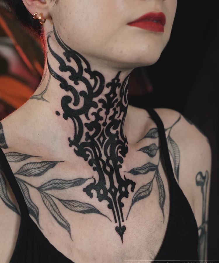 tatuagem-feminina-no-pescoco-22-1 A imagem mostra uma pessoa de frente com cabelo curto e batom vermelho. Ela tem uma tatuagem no pescoço que se estende até o peito, com um design intrincado e simétrico em preto. A pessoa usa brincos pequenos e uma blusa preta.