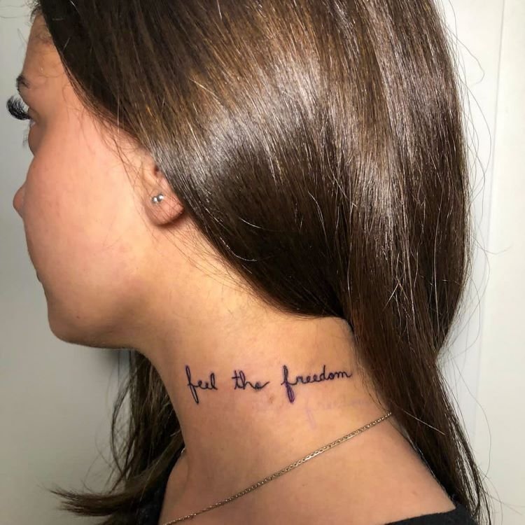 tatuagem-feminina-no-pescoco-24-1 A imagem mostra uma pessoa de perfil com cabelo liso e castanho. Ela tem uma tatuagem no pescoço com a frase "feel the freedom" escrita em letras cursivas pretas. A tatuagem está localizada na lateral do pescoço, logo abaixo da linha do cabelo, e a pessoa usa um brinco pequeno.