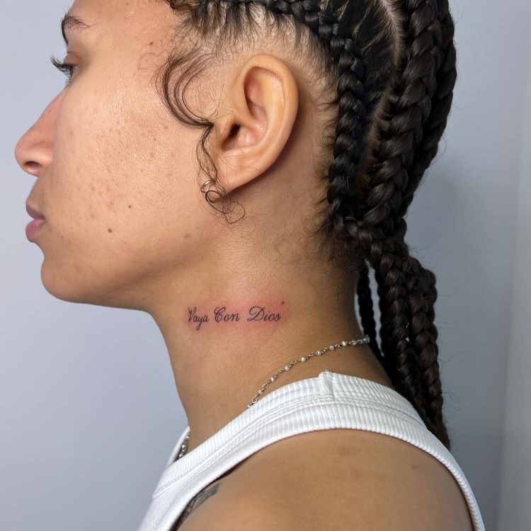 tatuagem-feminina-no-pescoco-30-1 A imagem mostra uma pessoa de perfil com cabelo trançado. Ela tem uma tatuagem no pescoço com a frase "Vaya Con Dios" escrita em letras cursivas pretas. A tatuagem está localizada na lateral do pescoço, logo abaixo da linha do maxilar. A pessoa usa uma corrente prateada.