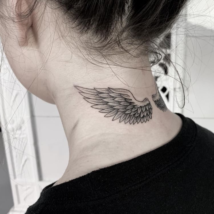 tatuagem-feminina-no-pescoco-34-1 A imagem mostra uma pessoa de costas com cabelo preso. Ela tem uma tatuagem no pescoço de asas de anjo estendendo-se horizontalmente, detalhadas em preto. A tatuagem está localizada na base do pescoço, logo abaixo da linha do cabelo, e é feita com traços finos e delicados.