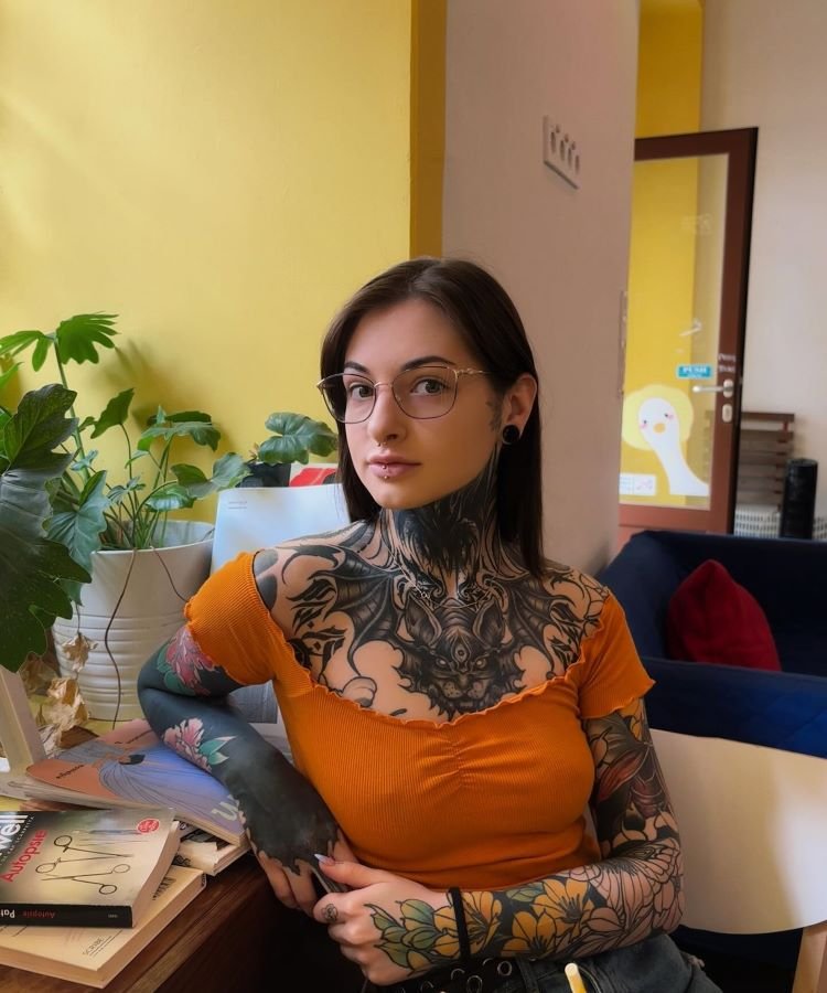 tatuagem-feminina-no-pescoco-37-1 A imagem mostra uma pessoa sentada, com cabelo castanho e óculos. Ela tem uma tatuagem no pescoço que se estende até o peito e ombros, com um design complexo em preto. A pessoa usa uma blusa laranja e está em um ambiente com plantas e livros ao redor.