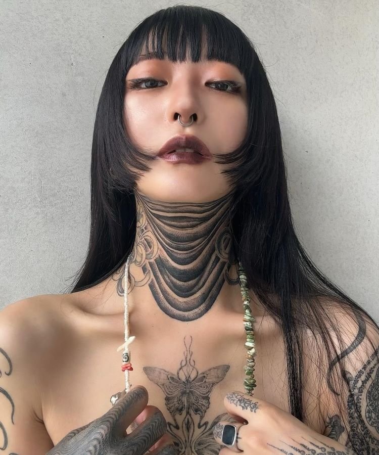 tatuagem-feminina-no-pescoco-39-1 A imagem mostra uma pessoa de frente com cabelo preto liso e franja. Ela tem uma tatuagem no pescoço com um design de várias faixas pretas curvas. A pessoa usa maquiagem marcante, um piercing no septo e segura um colar de contas brancas.