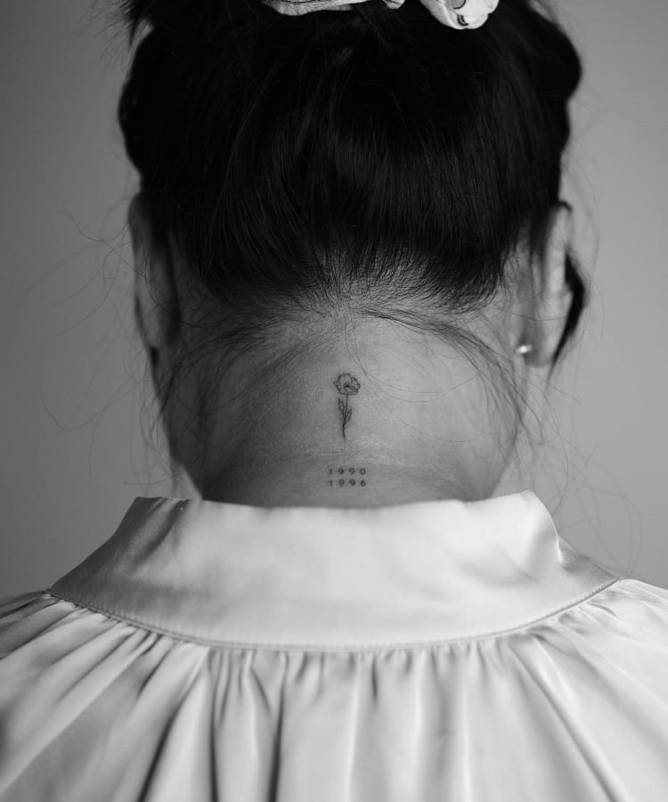 tatuagem-feminina-no-pescoco-41-1 A imagem mostra uma pessoa de costas com cabelo preso em um coque. Ela tem uma tatuagem no pescoço de uma pequena flor delicada com um caule fino e duas datas abaixo dela. A tatuagem está localizada na base do pescoço, centralizada.