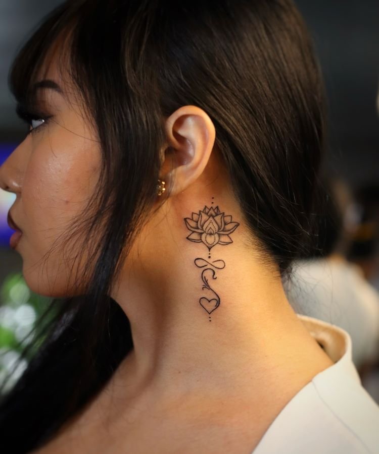 tatuagem-feminina-no-pescoco-5-1 A imagem mostra uma pessoa de perfil com cabelo escuro e liso. Ela tem uma tatuagem no pescoço de uma flor de lótus detalhada, com uma linha que desce até um coração. A tatuagem é delicada e em preto, localizada atrás da orelha e estendendo-se pela lateral do pescoço.