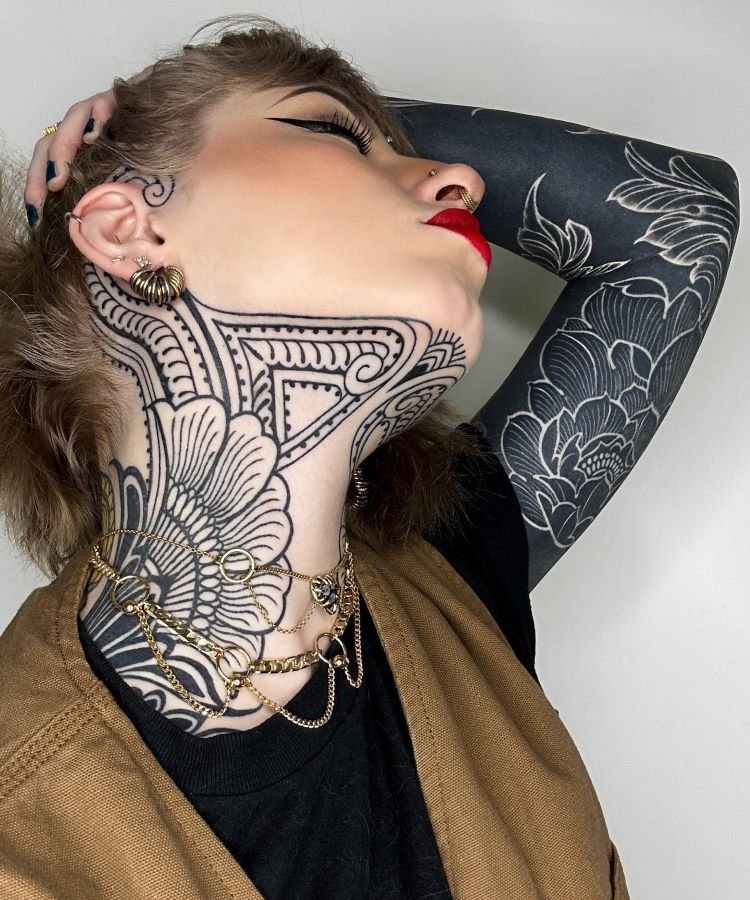 tatuagem-feminina-no-pescoco-9-1 A imagem mostra uma pessoa com a cabeça inclinada para trás, exibindo uma tatuagem no pescoço com desenhos detalhados em preto. Ela tem maquiagem marcante, batom vermelho e piercings. O braço direito também é tatuado com flores. Usa brincos dourados e colares de corrente.