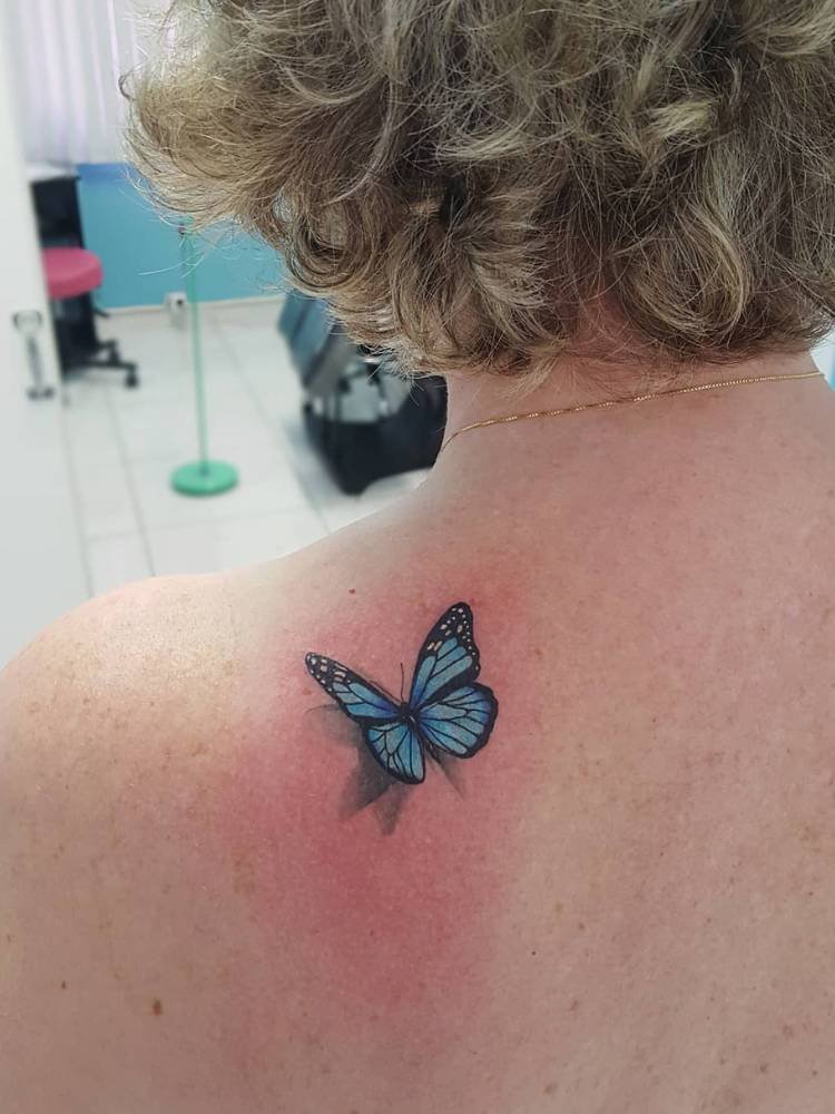 tatuagem-feminina-ombro-borboleta-azul-sombra-atras Mulher loira de cabelo curto com tatuagem de borboleta azul sombreada na parte de trás do ombro.